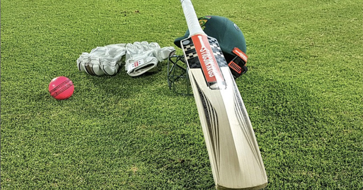 Cricket bat resting on a cricket helmet with cricket gloves nearby. All on a cricket field